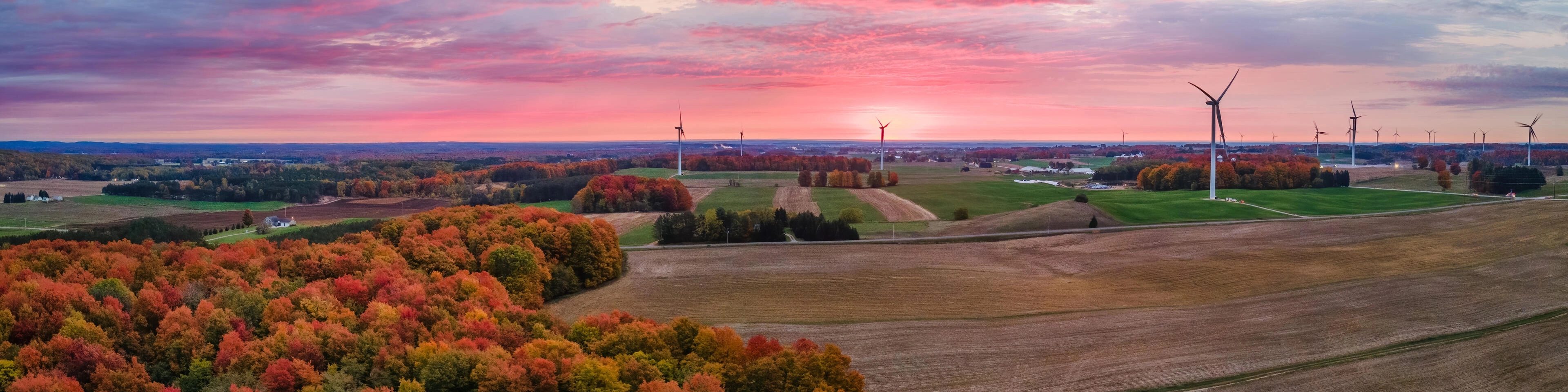 Panoramiczny widok z lotu ptaka na wiejski krajobraz o zachodzie słońca, przedstawiający lasy w jesiennych barwach, pola uprawne i liczne turbiny wiatrowe na tle różowo-niebieskiego nieba